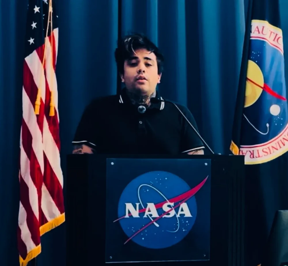 Eros Marcello speaking at a NASA podium, flanked by the US and NASA flags